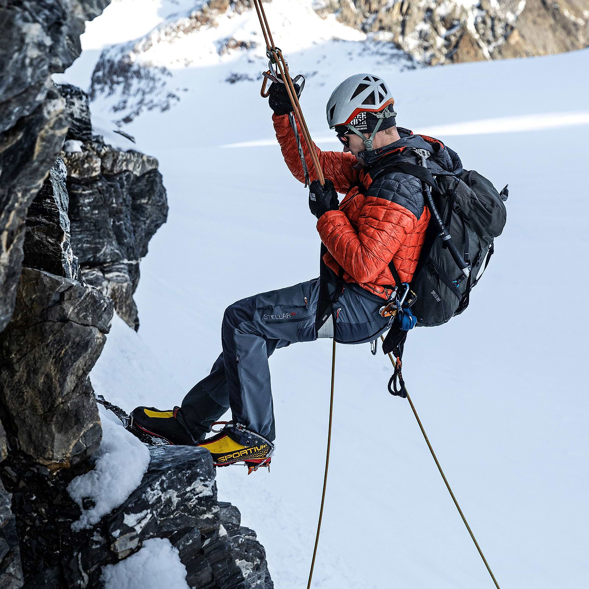 A mountaineering guide repelling down a rock face on Gross Spannort in Switzerland, wearing the Stellar Equipment Mens Hyperlight Down Hood in orange.