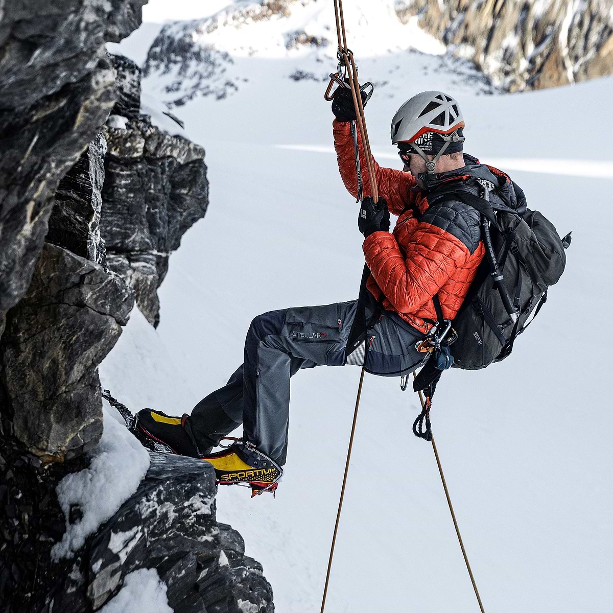 A mountaineering guide repelling down a rock face on Gross Spannort in Switzerland, wearing the Stellar Equipment Mens Hyperlight Down Hood in orange. The photo was taken by Joakim Åström.